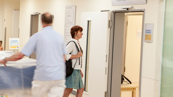 Eine Patientin geht in die Patientenaufnahme, ein Logistik-Mitarbeiter schiebt ein Patientenbett.