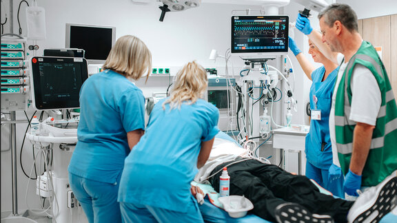 Vier medizinische Fachkräfte versorgen eine Person, die sich auf einer Krankenhausliege befindet, in einem modern ausgestatteten Behandlungsraum.