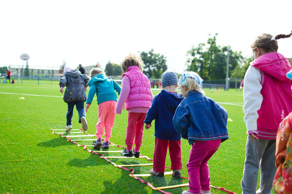 Kinder auf einer grünen Wiese in einer Reihe hintereinander machen ein Geschicklichkeits- und Hüpfspiel.