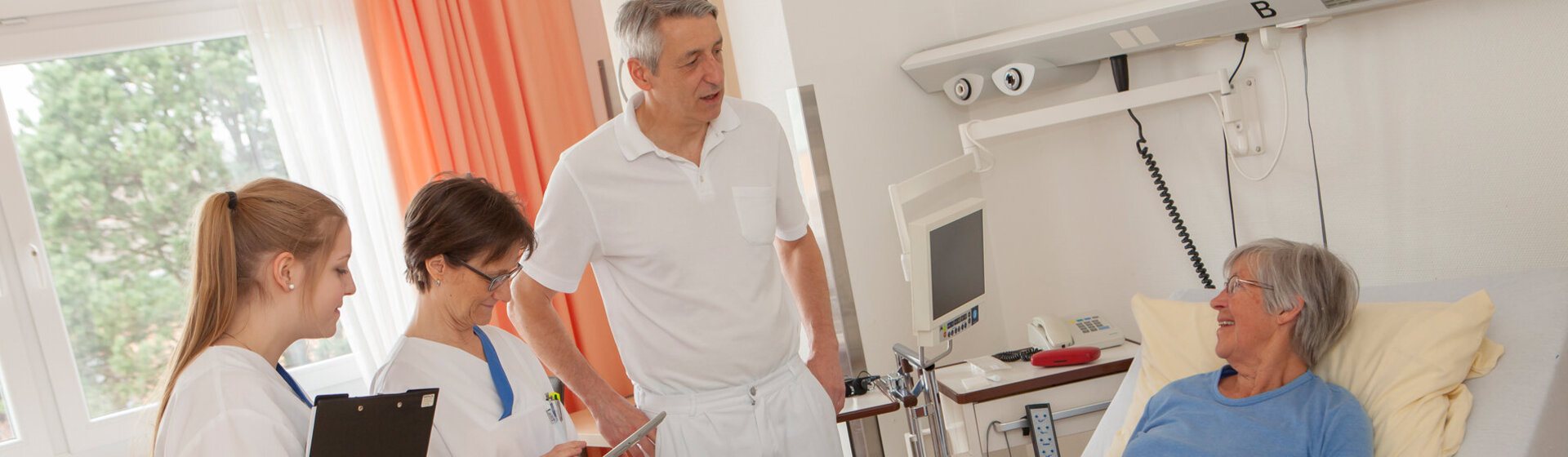 Prof. Dr. Michael Neipp und zwei medizinische Fachkräfte im Gespräch mit einer Patientin, die im Bett liegt.