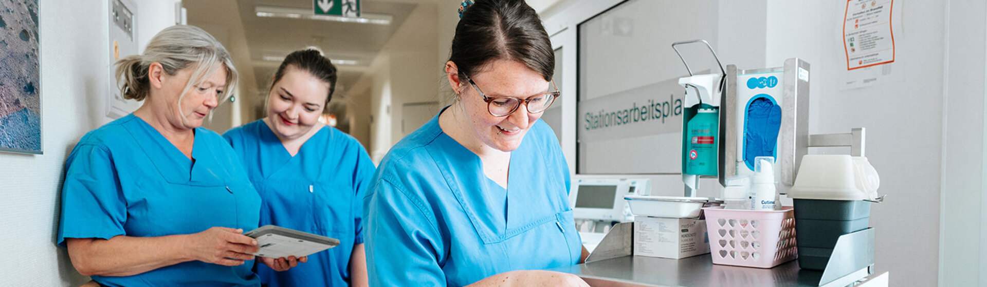 Drei Pflegefachkräfte stehen im Flur, zwei schauen auf ein Tablet, die dritte öffnet eine Schublade vom Medikamentenwagen.