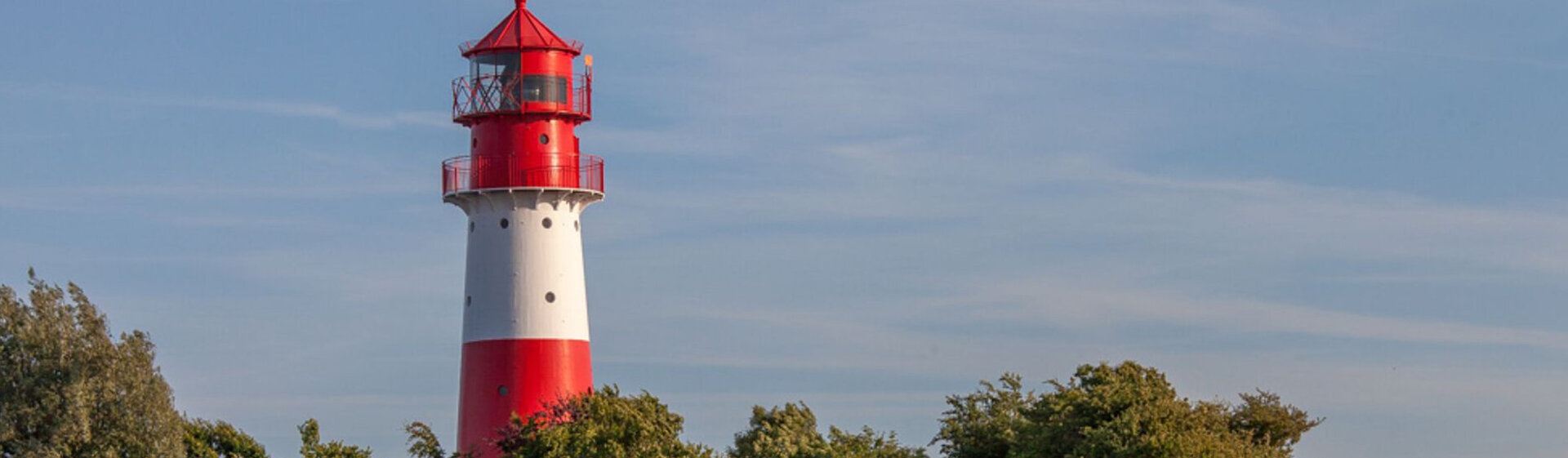Ein Leuchtturm steht hinter Baumkronen, im Hintergrund ist ein leicht bewölkter blauer Himmel erkennbar. Ein Leuchtturm steht hinter Baumkronen, im Hintergrund ist ein leicht bewölkter blauer Himmel erkennbar.