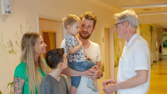 Chefarzt Dr. med. Georg Hillebrand (rechts) unterhält sich mit einem kleinen Patienten, der auf dem Arm des Vaters zusammen mit seiner Mutter und dem großen Bruder im Flur der Kinderklinik steht.