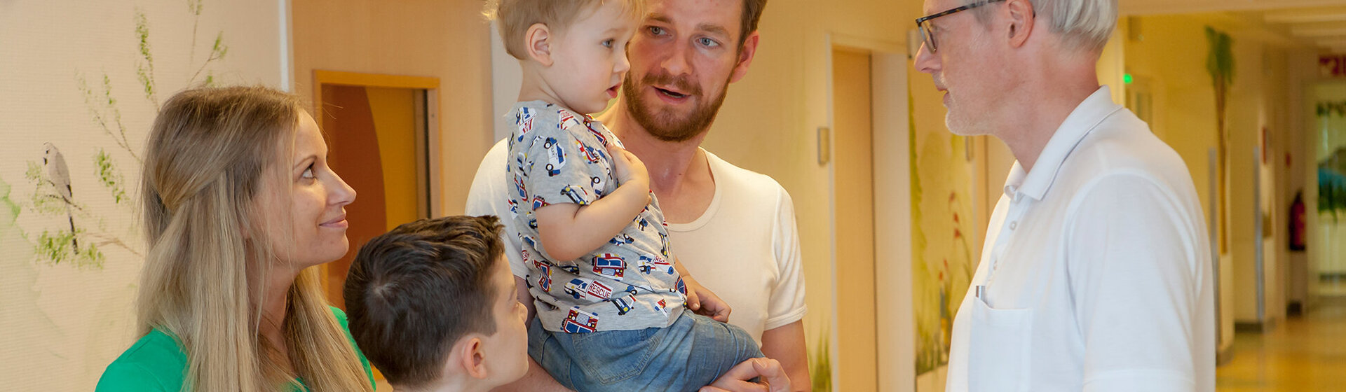 Chefarzt Dr. med. Georg Hillebrand (rechts) unterhält sich mit einem kleinen Patienten, der auf dem Arm des Vaters zusammen mit seiner Mutter und dem großen Bruder im Flur der Kinderklinik steht.