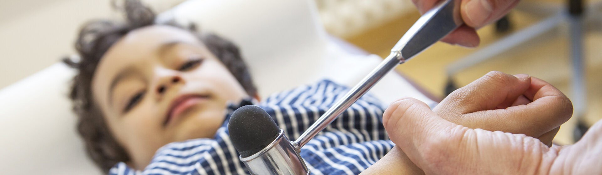 Ein Kind liegt auf einer Untersuchungsliege und wir von einem Arzt mit dem Reflexhammer untersucht.