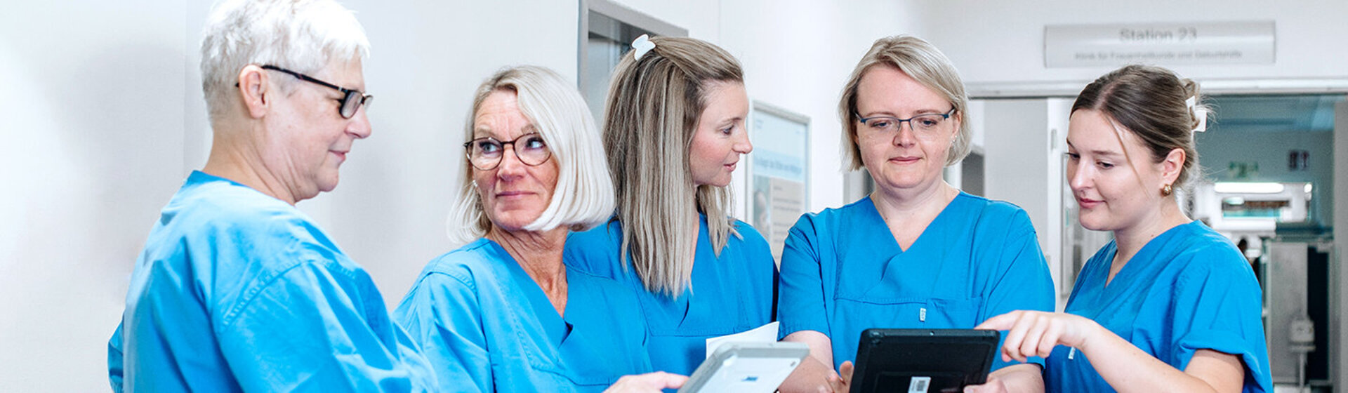 Fünf Pflegefachkräfte stehen am Stationstresen, schauen auf zwei Tablets und besprechen sich.