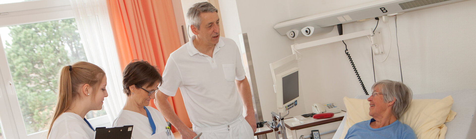 Chefarzt Prof. Dr. med. Michael Neipp und zwei medizinische Fachkräfte im Gespräch mit einer Patientin am PatientInnenbett.