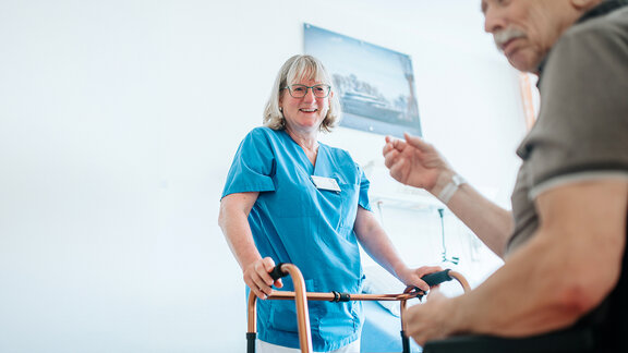 Patient sitzt im Rollstuhl, Pflegefachkraft lächelt ihn an und gibt ihm einen Gehwagen.