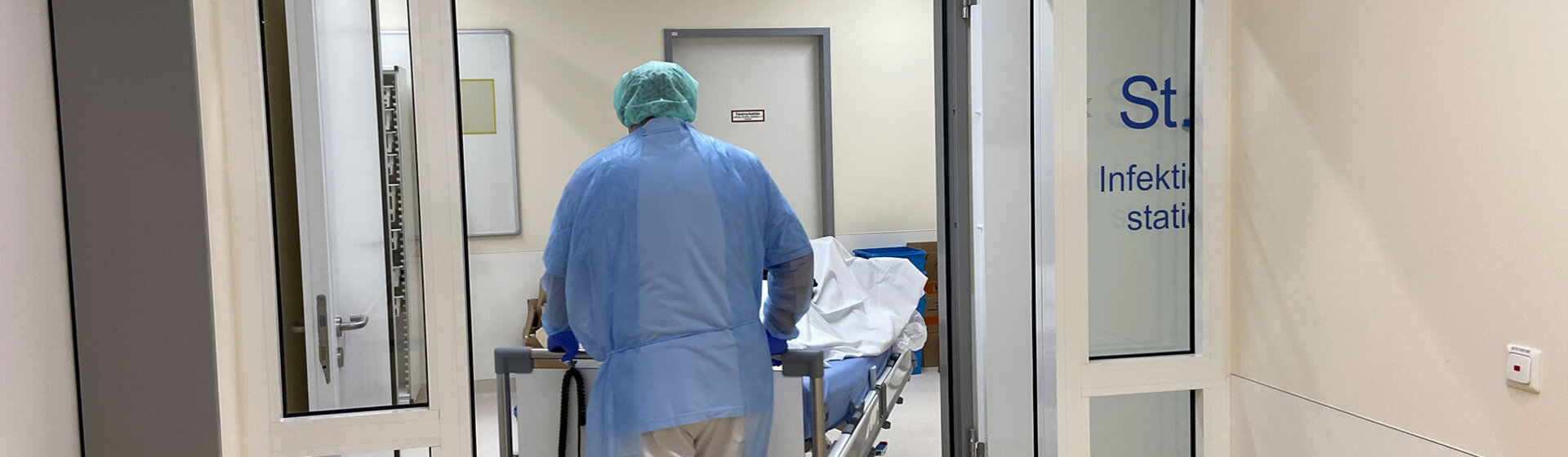 Ein Mitarbeiter mit Schutzkleidung schiebt ein PatientInnenbett durch die automatische Tür der Station 6. Infektionsstation Innenansicht