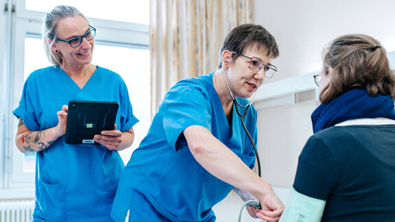 Zwei Pflegefachkräfte stehen am PatientInnenbett, die Patientin sitzt im Bett, eine Pflegefachkraft misst Blutdruck.