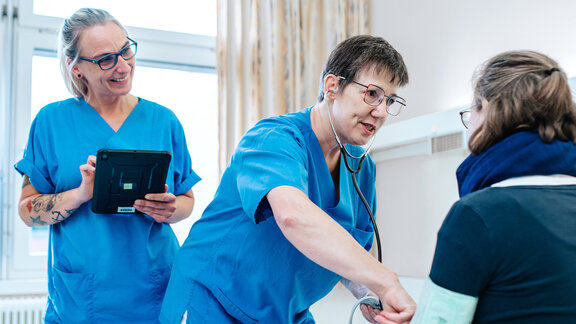 Zwei Pflegefachkräfte stehen am PatientInnenbett, die Patientin sitzt im Bett, eine Pflegefachkraft misst Blutdruck.