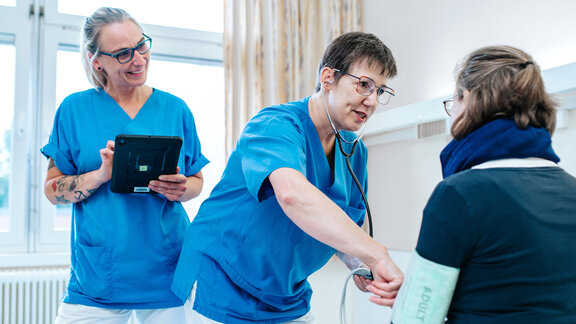 Zwei Pflegefachkräfte stehen am PatientInnenbett, die Patientin sitzt im Bett, eine Pflegefachkraft misst Blutdruck.
