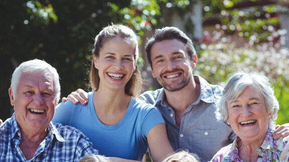 Eine fröhliche Familie bestehend aus Großeltern, Eltern und Kindern sitzen zusammen im Grünen und lächeln in die Kamera.