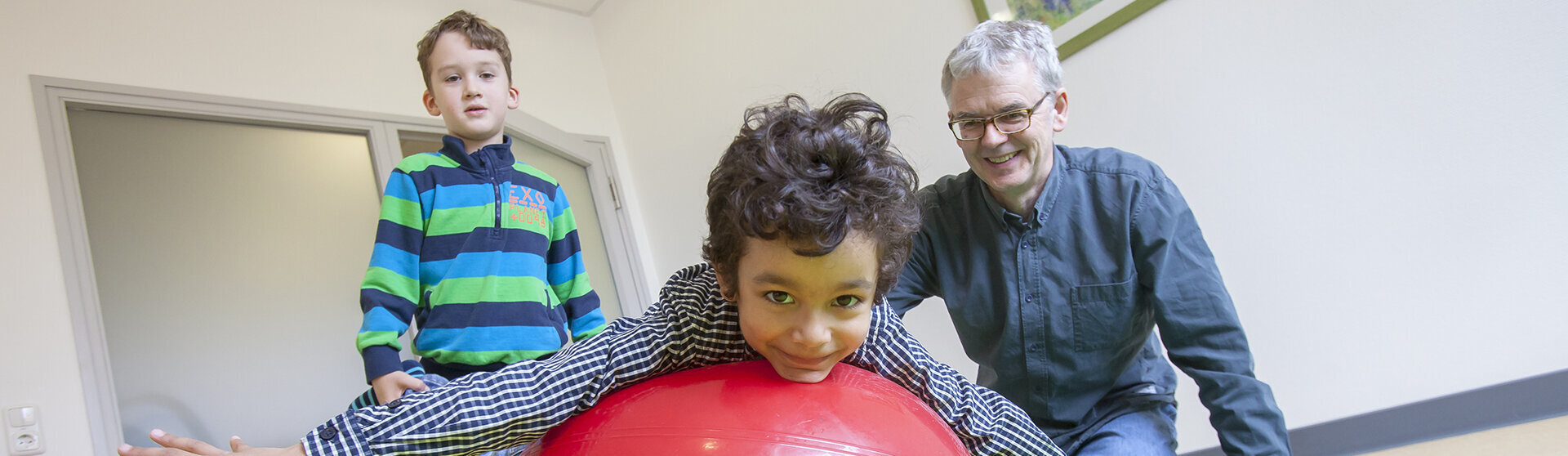 Physiotherapeut Sven Olde mit zwei Kindern in der Therapiesituation auf einer Gymnastikmatte, ein Kind liegt bäuchlings auf einem roten Gymnastikball, das zweite Kind hält die Füße des ersten Kindes. Physiotherapeut Sven Olde mit zwei Kindern in der Therapiesituation auf einer Gymnastikmatte, ein Kind liegt bäuchlings auf einem roten Gymnastikball, das zweite Kind hält die Füße des ersten Kindes.