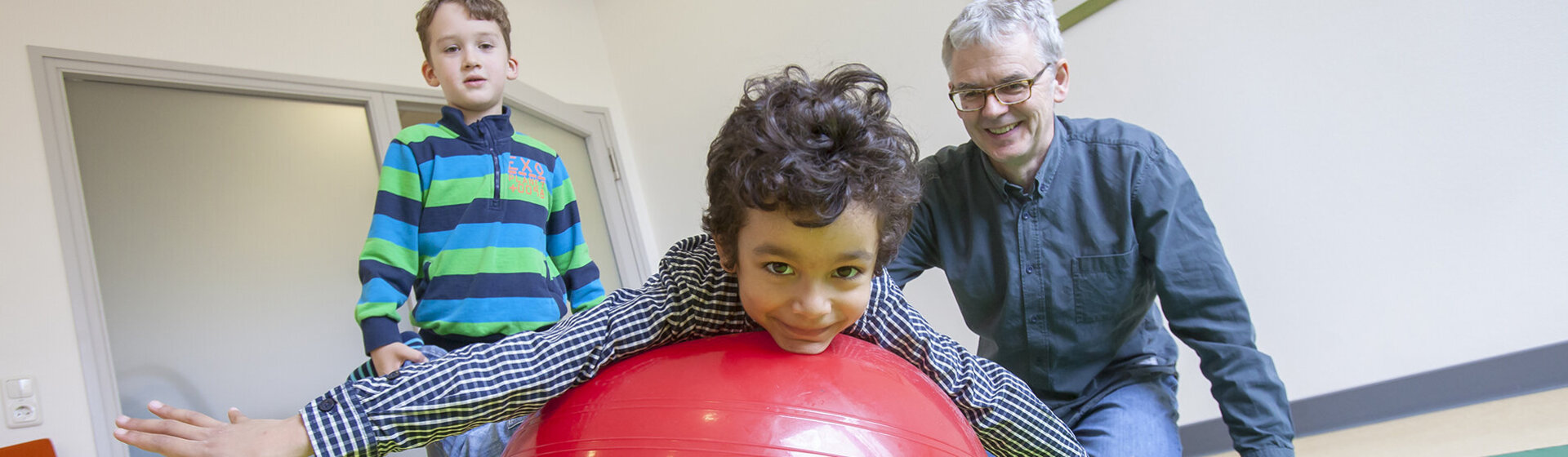 Physiotherapeut Sven Olde mit zwei Kindern in der Therapiesituation auf einer Gymnastikmatte, ein Kind liegt bäuchlings auf einem roten Gymnastikball, das zweite Kind hält die Füße des ersten Kindes.