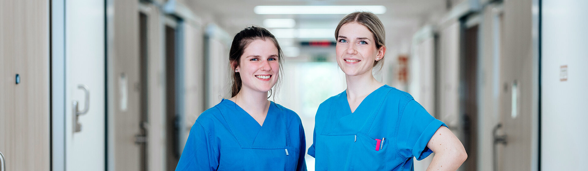 Zwei lächelnde Pflegefachkräfte, im Hintergrund der Krankenhausflur
