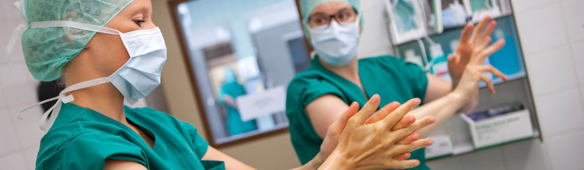 Zwei medizinische OP-Fachkräfte in grüner OP-Kleidung bereiten sich auf eine OP vor und desinfizieren Hände und Arme.