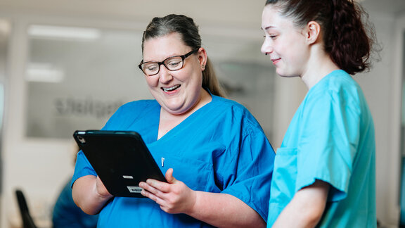 Zwei Pflegefachkräfte schauen lächelnd auf ein Tablet.