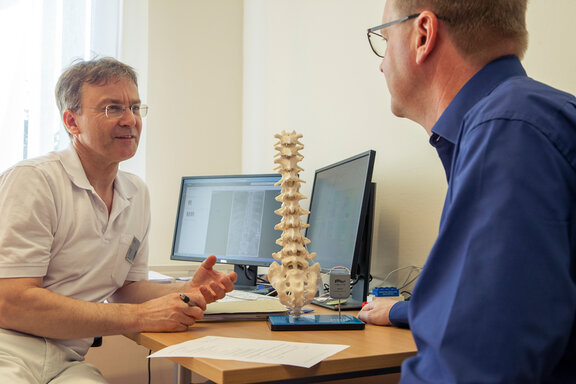 Dr. med. Wolfram Kluge klärt einen Patienten anhand eines Wirbelsäulenmodells über die SCS-Rückemankstimulation auf.