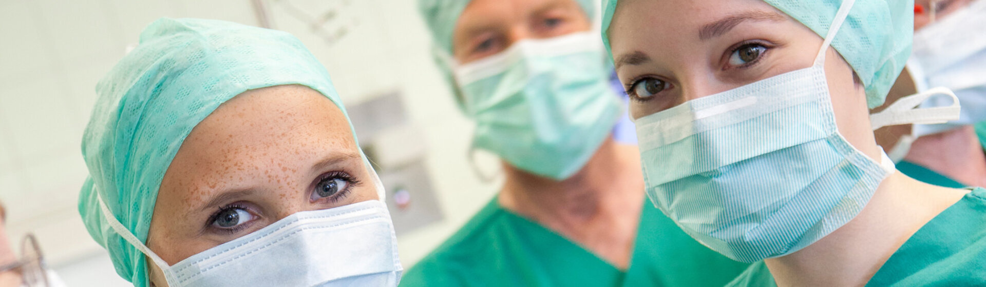 Drei Mitarbeitende in OP-Kleidung mit Mundschutz und Haube lächeln ins Bild.