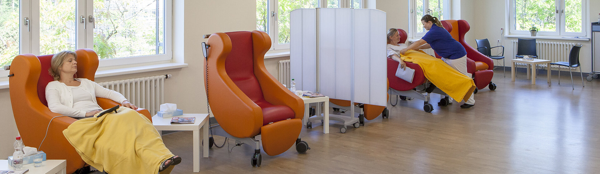 Heller Aufwachraum mit rot-orangefarbenen Aufwachsesseln in denen PatientInnen sitzen und von einer Pflegefachkraft betreut werden Heller Aufwachraum mit rot-orangefarbenen Aufwachsesseln in denen PatientInnen sitzen und von einer Pflegefachkraft betreut werden