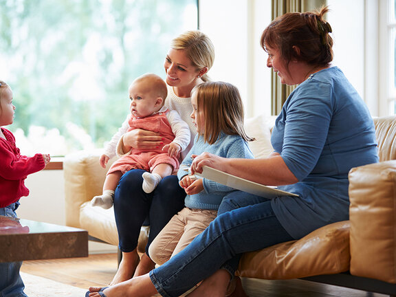 Zwei Frauen sitzen mit drei Kindern unterschiedlichen Alters in einem hellen Wohnzimmer