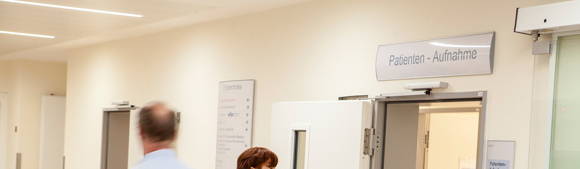 Eine Patientin geht in die Patientenaufnahme, ein Logistik-Mitarbeiter schiebt ein Patientenbett.