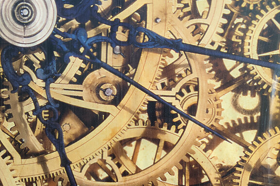 Zahnräder einer Uhr greifen ineinander, darüber sind blaue Zeiger abgebildet.