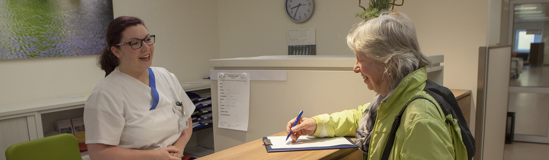 Pflegefachkraft lächelt Patientin an, die am Anmeldetresen steht und etwas ausfüllt.