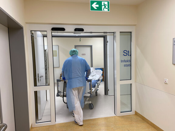 Ein Mitarbeiter mit Schutzkleidung schiebt ein PatientInnenbett durch die automatische Tür der Station 6.