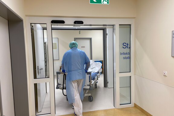 Ein Mitarbeiter mit Schutzkleidung schiebt ein PatientInnenbett durch die automatische Tür der Station 6.