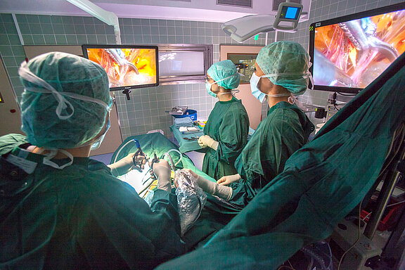 Operationssituation eines Minimal-invasiven Eingriffs, Operateur und medizinische Fachkräfte blicken auf den OP-Monitor.