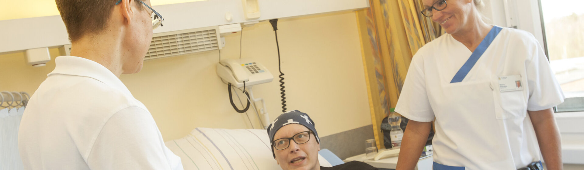 Patientin liegt im Bett, zwei Pflegefachkräfte stehen neben dem Bett und lächeln sie an.