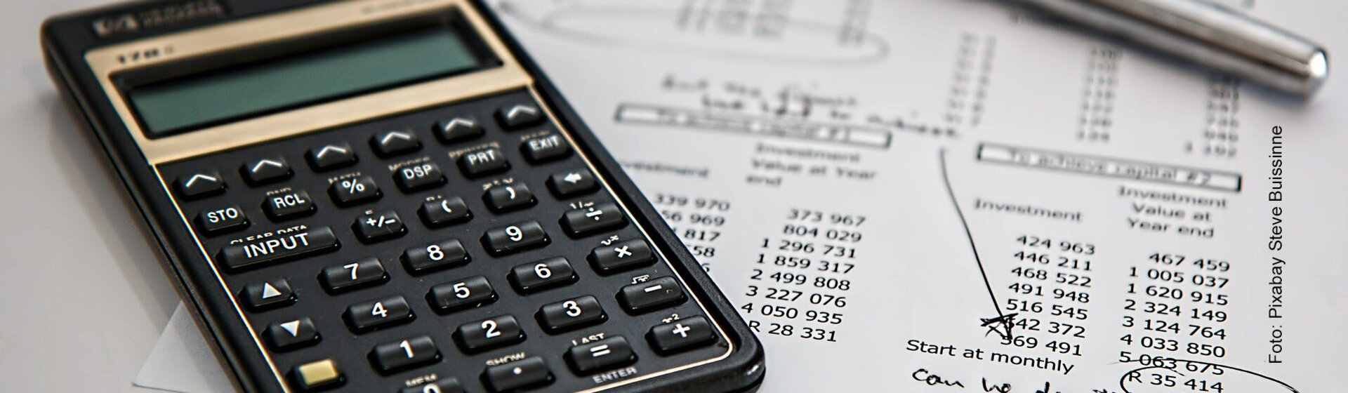 Taschenrechner, Kugelschreiber und Zettel mit Zahlen liegen auf einem Tisch.