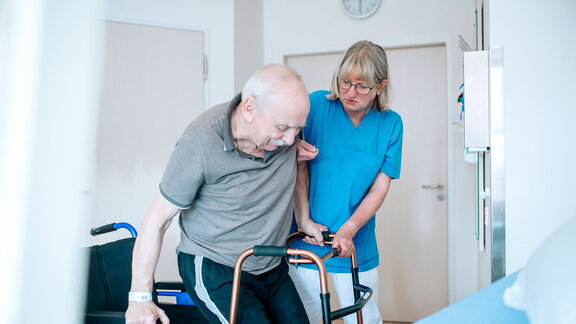 Eine Pflegefachkraft hilft einem Patienten aus dem Rollstuhl aufzustehen.